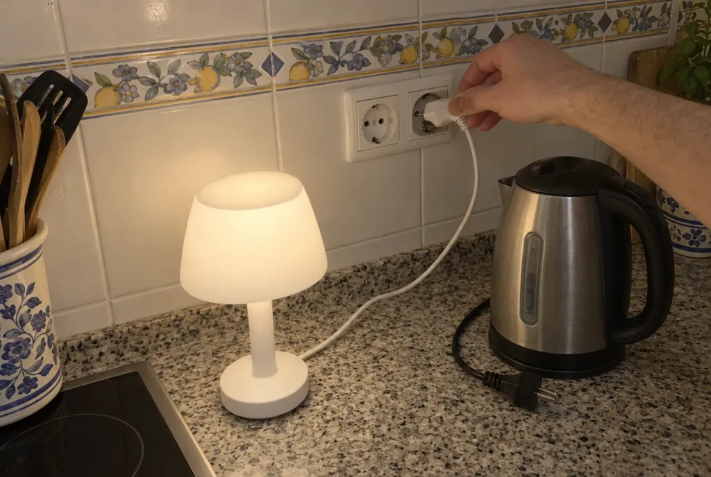 Mano enchufando una lámpara pequeña en un enchufe doble de la cocina, sobre la encimera, junto a un hervidor